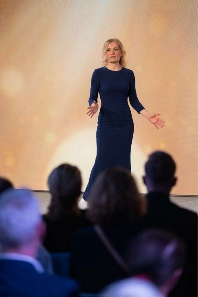 Katharina Ivanenko spricht auf einer Bühne mit ausgebreiteten Armen vor Publikum, hinter ihr ein warm goldener Hintergrund